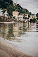 Fototapeta na wymiar Beautiful view on old buildings on lake in Stresa city, Italy. Architecture and shore on Lago Maggiore in sunny day on background of palms and mountains. Summer vacation in Europe