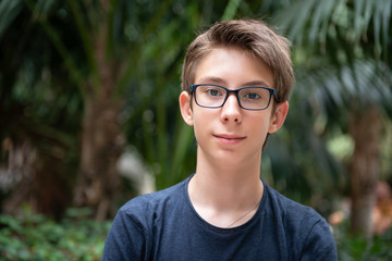 Young boy posing in summer park with palm trees. Cute spectacled smiling happy teen boy 13 years...
