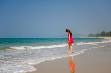 Young slender long-haired brunette in red dress walks barefoot on tropical beach along the surf against the sea and blue sky on a hot day. In the distance a green hill and people on the shore.