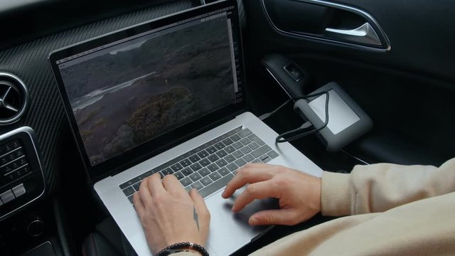 Closeup on trendy and hipster creative professional with tattooed hands work on laptop remotely from passenger seat in car during travel or roadtrip. Watches videos on computer and makes to do list
