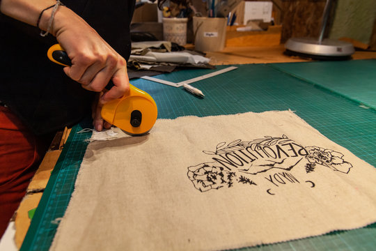 Fashion designer is cutting patches. A close up and side view on the hands of a young caucasian eco-designer using a rotary cutter to trim a piece of beige fabric with floral print pattern.