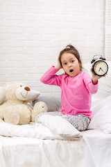 Surprised cute little child girl in pyjamas with clock in bed. good morning