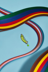 Closeup shot of shiny breastpin in view of peapod with pendant in view of pea, isolated asymmetrically on light blue background in midst of multicolored wavy placed ribbons. Voguish fashion item.