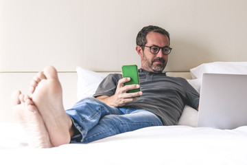 Businessman communicate with remote colleagues during busines trips around the world sitting on bed after work. Casual dressed man using laptop and smart phone in an hotel room. New technolgy concept.