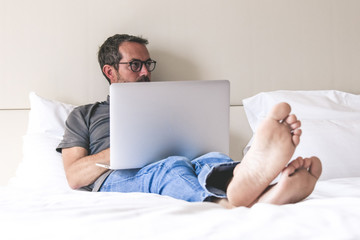 Barefoot young man sitting on a bed relaxing after work using laptop. Leisure time for a man alone in an hotel room watching new trends video online. New technology relax and social media concept