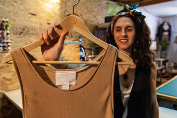 Fashion designer is showing a unisex top. Happy dressmaker is seen holding beige clothing on wooden hanger inside workshop. Finished product of creative crafts person.