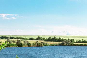 Summer landscape with a beautiful blue lake and green fields against a blue sky. Hiking, fishing on the pond. Journey to the river on a warm day