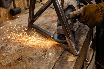 Metalworker uses disc grinder on steel. A closeup view of hot sparks flying from the abrasive disc of an angle grinder whilst a blacksmith polishes a metal frame. - obrazy, fototapety, plakaty
