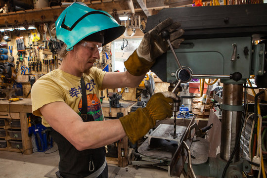 Man using pillar drill inside workshop. A metalworker operates the lever of a bench drill to pierce a hole through tough metal. Heavy duty machine inside the workshop of a blacksmith.