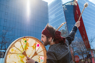 Sacred man meets urban environment. A closeup view of a spiritual guy playing a traditional drum downtown, seeks inspiration from city surroundings. Blurred skyscrapers in background.