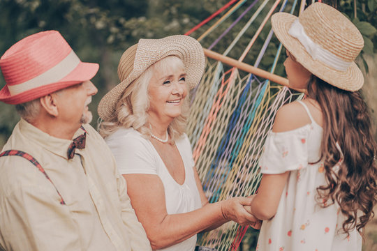 Old Couple In The Garden Sitting On Hammock Back View. Grandfather In Red Hat Loves Grandmother. Happy Old Age. Young Granddaughter Runs To Them