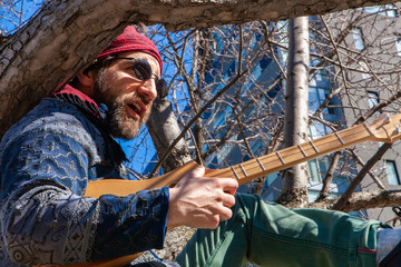 Creative man sits in tree by skyscraper. A bearded shaman is viewed close-up, holding an acoustic guitar and sitting in a tree on a city street, commercial buildings are seen in background.