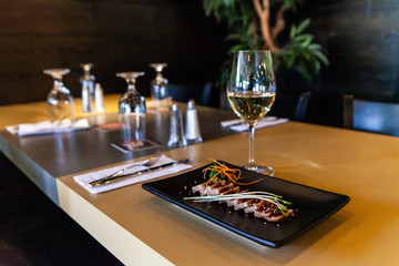 Raw tuna dish served in restaurant. A closeup view of tuna tataki, a briefly seared fish dish marinated in vinegar, served with white wine on a bistro table, with blurry glasses and tableware.