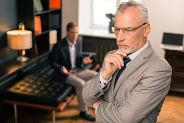 Serious pensive grey-haired psychoanalyst standing in his office