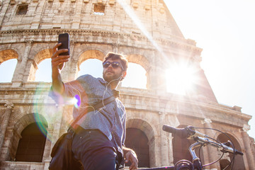 Young man with bike taking selfies at colosseum in Rome. Blue shirt, sunglasses on sunny summer day