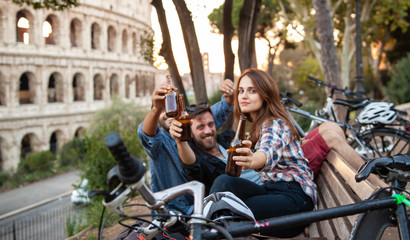 Three happy young friends tourists on a bench at Colosseum in Rome having fun drinking and toasting...