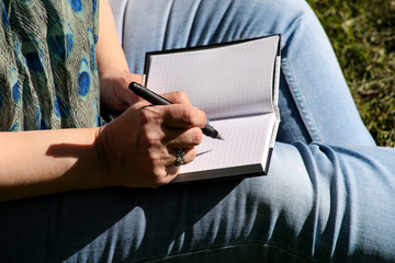 Woman and student sitting on grass, takes notes in notebook, learning and writes thoughts, writes book, she is preparing his final exams doing homework and thinking, writing something in his notebook.