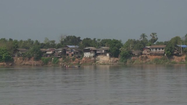 A daylight long shot of a small pump boat carrying local passengers and passing through the murky waters of the river near a residential coastline.