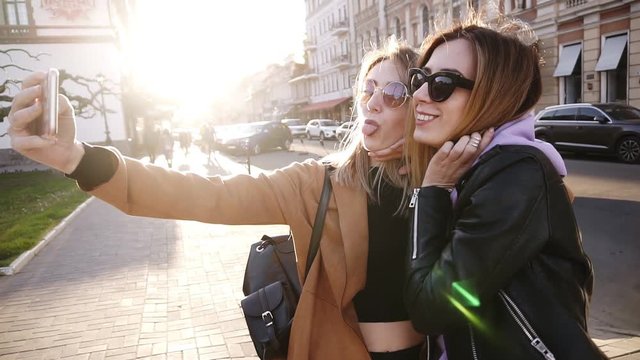 Two young caucasian fashion women in sunglasses posing for selfie and laughing with tonque. Lifestyle portrait on street city background