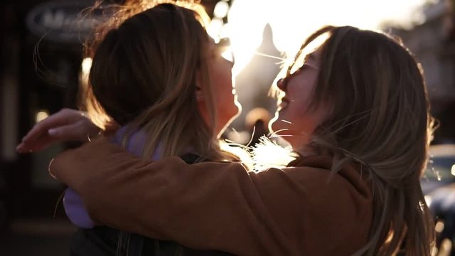 Two happy young girls hug each other. Females embracing, laughing and excited. Woman friendship, walk in the city outdoors. City view, sunlight on the background
