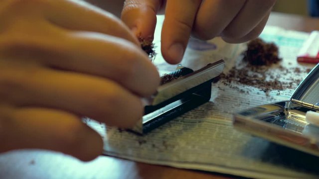 A young man makes a handmade cigarette filling paper with shredded tobacco using a rolling machine. On the table is a newspaper and an open cigarette case.