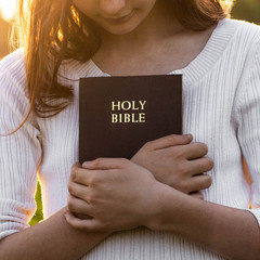 Christian teenage girl holds bible in her hands. Reading the Holy Bible in a field during beautiful...