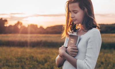 Christian teenage girl holds bible in her hands. Reading the Holy Bible in a field during beautiful sunset. Concept for faith, spirituality and religion. Peace, hope - obrazy, fototapety, plakaty