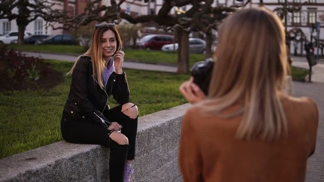 Footage from the shoulder - professional female photographer takes pictures of a girl model her friend. Attractive long haired woman in black casual clothes posing for the camera, sitting on the