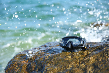 Black diving mask on a rocky seashore close up against the background of ocean waves crashing on the stones. Wet diving mask on the stone in the sea.