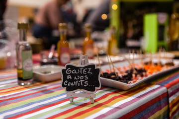 Seasonal products sold at Xmas market. A small French sign is viewed close-up, saying taste our confits, blurry cocktail sticks with preserved meats are seen in the background during a festive fair.
