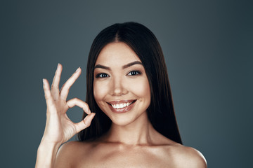Everything is OK. Beautiful young Asian woman looking at camera and smiling while standing against...