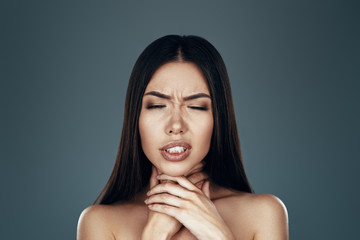 Sore throat. Frustrated young Asian woman touching her throat while standing against grey background
