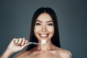 Obraz premium Taking good care of her teeth. Beautiful young Asian woman smiling and looking at camera while standing against grey background