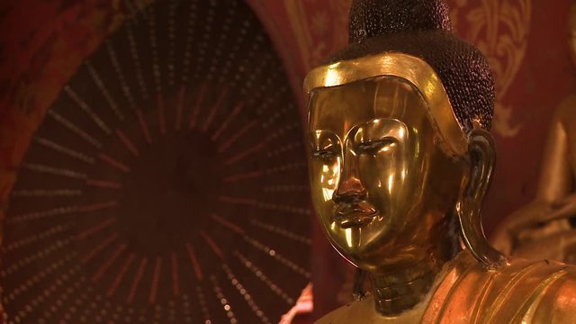 A steady closeup shot of the smooth face of a golden Buddha statue against a red painted wall background inside a Buddhist museum, taken in a daylight setting.