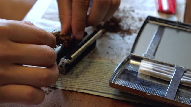 A young man makes a handmade cigarette filling paper with shredded tobacco using a rolling machine. On the table is a newspaper and an open cigarette case.