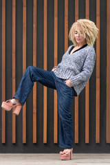 Hipster image. European girl blonde hairstyle afro curls originally posing in full growth in gray knitted sweater, free blue jeans and chunky hell.