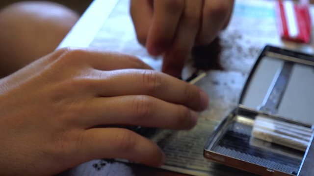 A young man makes a handmade cigarette filling paper with shredded tobacco using a rolling machine. On the table is a newspaper and an open cigarette case.