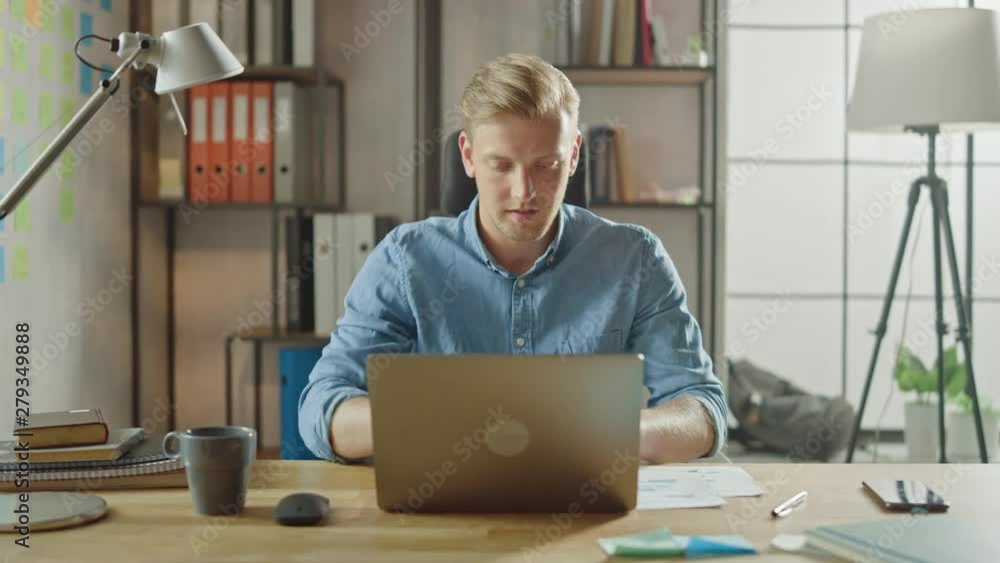 Poster handsome young entrepreneur enters his office sits at the desk opens laptop and starts working on th