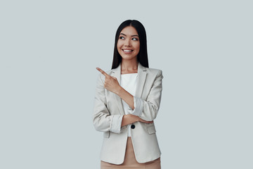 And here comes... Attractive young Asian woman looking away and pointing copy space while standing against grey background
