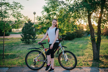 Active life. Sporty young blonde girl in sports clothes with a backpack rides a bike and smiles at the camera in a green Park on a summer evening at sunset. Sport and healthy lifestyle