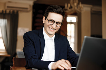 Portrait of a attractive confident businessman dressed in suit and wearing glasses looking at camera smiling while working at his laptop in his loft office.