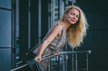 Fototapeta na wymiar Street fashion. A beautiful tall slim blond woman with magnificent hair in a trendy long dress with a reptile print (snake) clings to a pyrill near a modern gray building.