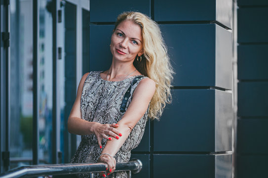 Street fashion. A beautiful tall slim blond woman with magnificent hair in a trendy long dress with a reptile print (snake) clings to a pyrill near a modern gray building.