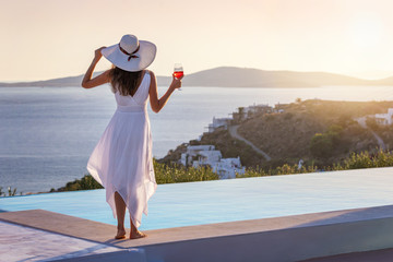 Attraktive Frau in weißem Sommerkleid genießt den Sonnenuntergang am Pool mit einem Aperitif in...