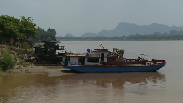A hand held, long shot of two small, old boats anchored near the shore where the sand meets the green and lively vegetation.