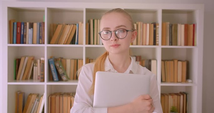 Closeup portrait of young pretty caucasian blonde female student in glasses holding a laptop looking at camera standing indoors in the library