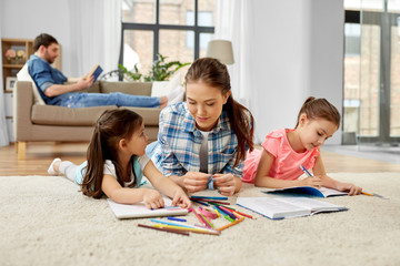 family, motherhood and leisure concept - mother spending time with her little daughters drawing and helping with homework lying on floor at home