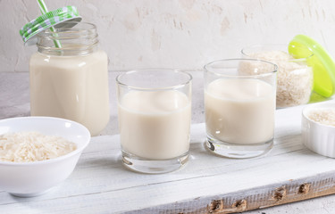 Drinking glasses, glass mason mug with  rice milk, bowl and jar with rise seeds on wooden board on white background. 