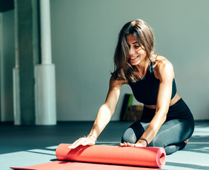 Smiling woman in sport clothes rolling mat in yoga studio