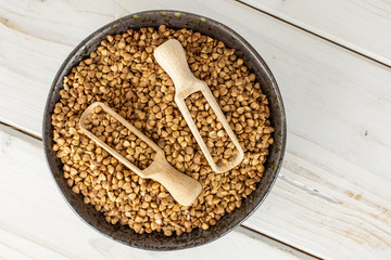 Lot of whole raw brown buckwheat grain in a grey ceramic bowl with wooden scoop flatlay on white wood